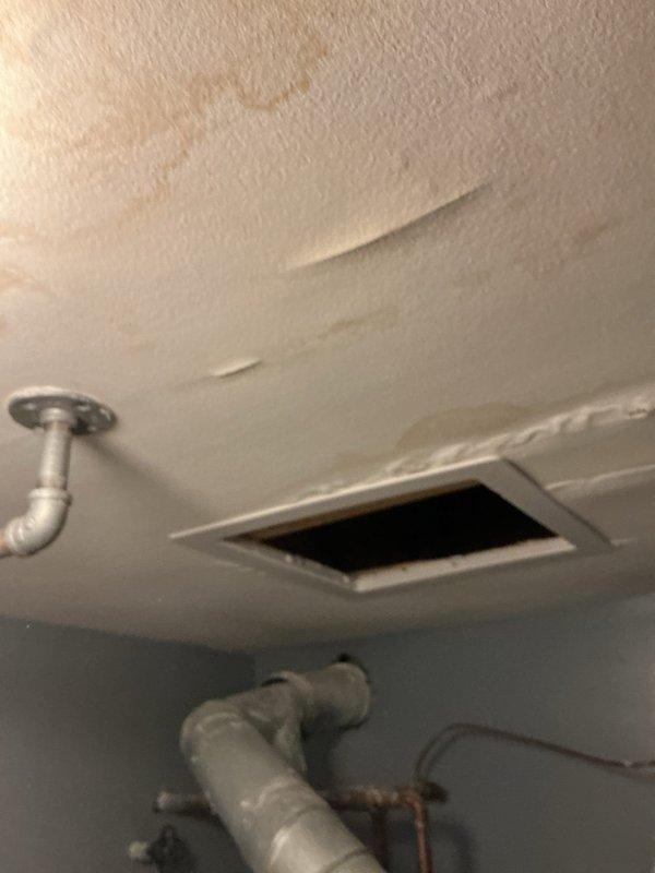 Responded to water leak investigation in laundry room area where customer reported slow leak appearing around ceiling light fixture, suspected to originate from unit above. Performed visual inspection of affected ceiling area, documenting extensive water staining and discoloration on textured ceiling surface near plumbing penetrations and vent opening. Examined exposed piping infrastructure including copper lines, PVC components, and metal fixtures in utility space to identify potential leak source. Based on staining patterns and pipe locations, determined leak likely originates from plumbing system in upper unit requiring coordination with above-unit access for definitive source identification and repair.