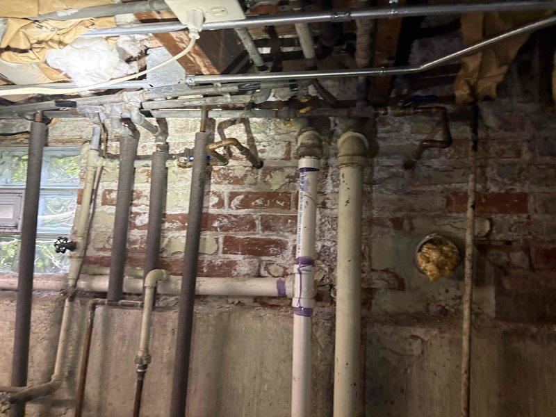 Completed comprehensive plumbing estimate for piping system replacement in utility area featuring deteriorated copper supply lines with extensive corrosion and mineral deposit buildup, aging PVC drain and waste lines with visible purple primer at connections, and compromised infrastructure along exposed brick foundation wall. Documented existing conditions including corroded brass fittings, valves requiring replacement, and multiple pipe runs servicing the mechanical systems in preparation for complete repiping specifications.