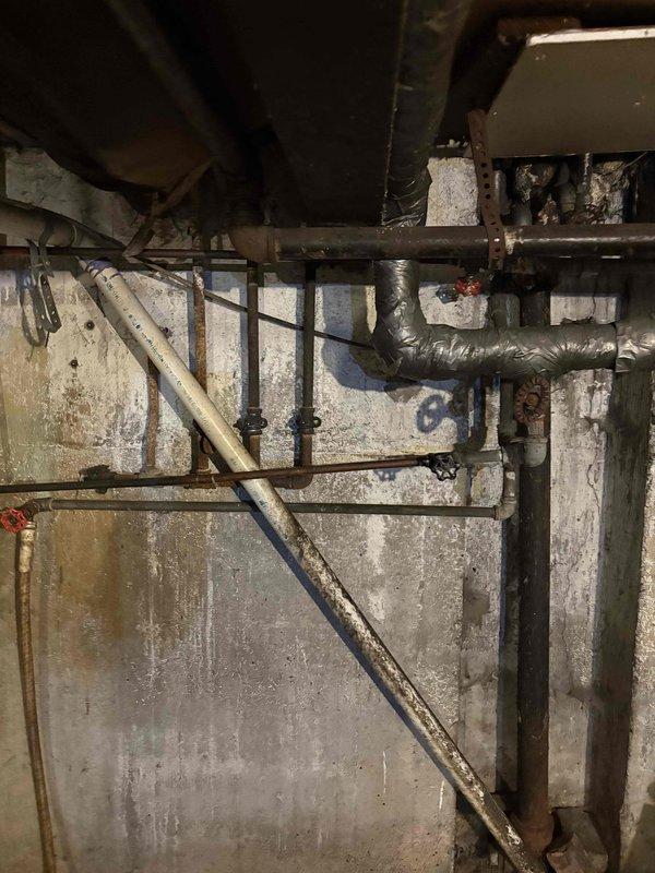 Conducted comprehensive plumbing assessment of aging basement mechanical infrastructure, documenting extensive deterioration of existing piping system including corroded metal components, compromised pipe joints, and weathered fixtures. Photographed moisture-damaged concrete block walls exhibiting efflorescence and mold growth, along with multiple horizontal and vertical pipe runs showing significant corrosion requiring replacement or repair recommendations for copper, galvanized, and flexible ductwork connections.