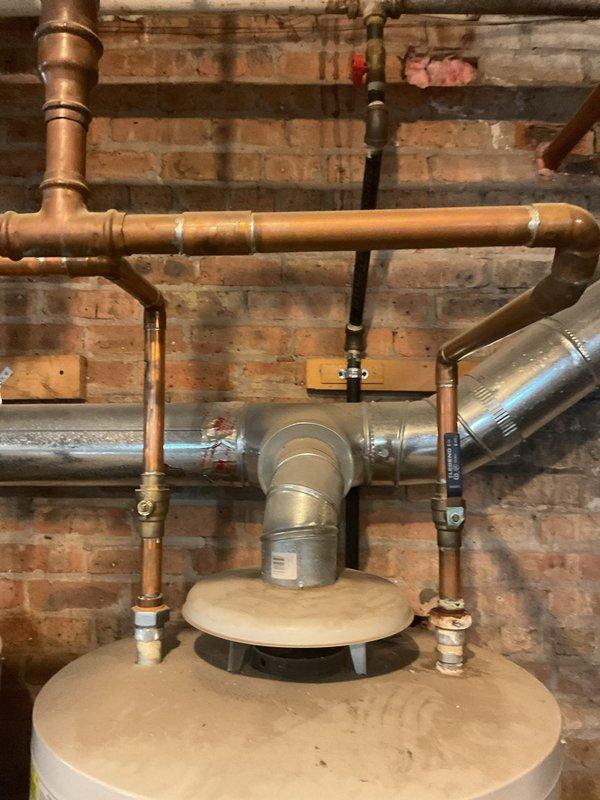Conducted on-site assessment for water heater replacement in basement utility area. Evaluated existing cylindrical storage-type water heater installation including all associated copper supply lines, gas piping with shut-off valves, and metal exhaust venting system against exposed brick wall. Documented current plumbing configuration showing multiple connection points, T-fittings, and routing of hot and cold water lines. Assessed gas line assembly with red-handle valve and various pipe fittings for compatibility with new unit installation. Noted floor conditions, moisture concerns, and workspace accessibility for equipment removal and replacement. Photographed complete system layout to prepare updated estimate for building water heater replacement project.