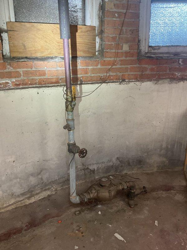 Conducted on-site assessment for water heater replacement in basement utility area. Evaluated existing cylindrical storage-type water heater installation including all associated copper supply lines, gas piping with shut-off valves, and metal exhaust venting system against exposed brick wall. Documented current plumbing configuration showing multiple connection points, T-fittings, and routing of hot and cold water lines. Assessed gas line assembly with red-handle valve and various pipe fittings for compatibility with new unit installation. Noted floor conditions, moisture concerns, and workspace accessibility for equipment removal and replacement. Photographed complete system layout to prepare updated estimate for building water heater replacement project.