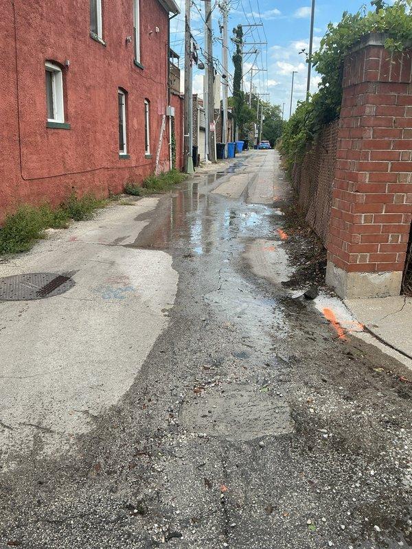 Responded to service call for water main leak causing groundwater to surface in residential alleyway and driveway area. Upon arrival, observed extensive standing water and wet pavement throughout alley, with water pooling near utility infrastructure and residential structures. Conducted visual inspection of affected areas, including concrete driveway with existing deterioration, cracking, and previous repair patches. Located circular utility cover in driveway surface near leak origin point. Documented existing utility markings and water damage patterns on concrete surfaces. Photographed wet conditions in alleyway extending between brick buildings, standing water accumulation on mixed concrete and asphalt surfaces, and compromised driveway areas adjacent to garage and brick wall structures. Prepared comprehensive estimate for water main repair work, including excavation requirements, line replacement specifications, and surface restoration needs for both alleyway and driveway areas affe