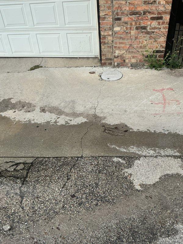 Responded to service call for water main leak causing groundwater to surface in residential alleyway and driveway area. Upon arrival, observed extensive standing water and wet pavement throughout alley, with water pooling near utility infrastructure and residential structures. Conducted visual inspection of affected areas, including concrete driveway with existing deterioration, cracking, and previous repair patches. Located circular utility cover in driveway surface near leak origin point. Documented existing utility markings and water damage patterns on concrete surfaces. Photographed wet conditions in alleyway extending between brick buildings, standing water accumulation on mixed concrete and asphalt surfaces, and compromised driveway areas adjacent to garage and brick wall structures. Prepared comprehensive estimate for water main repair work, including excavation requirements, line replacement specifications, and surface restoration needs for both alleyway and driveway areas affe