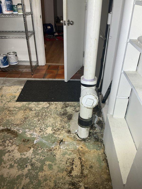 Responded to clogged drain service call in utility room area. Upon arrival, assessed drainage system consisting of white PVC vertical stack with T-junction fitting and black rubber couplings. Cleared obstruction from drain line to restore proper flow and functionality. Documented existing water damage on concrete floor surface, indicating previous drainage issues in the area.