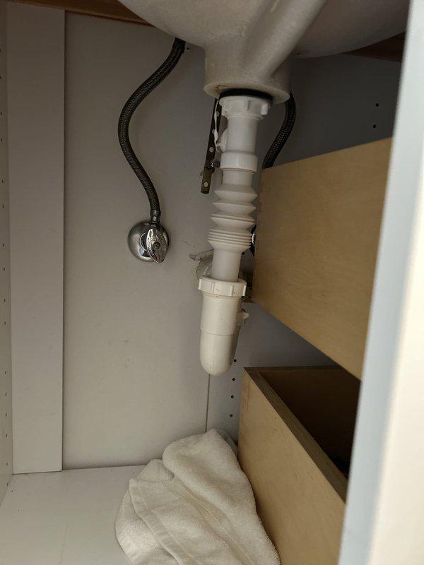 Responded to service call for multiple plumbing issues including dishwasher leaking from bottom due to suspected clogged valve, bathroom sink with minor leak at drain assembly, and toilet replacement with customer-supplied fixture. Inspected bathroom sink P-trap and drain connections beneath cabinet, identifying source of leak at flexible accordion-style tubing connection. Documented existing plumbing configuration including white PVC P-trap assembly, chrome shut-off valves, and black braided supply lines for future reference during repairs.