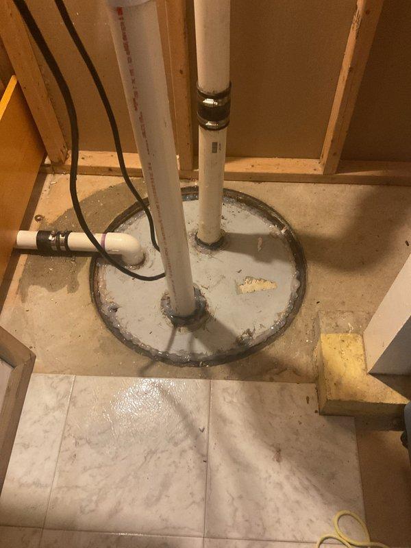 Responded to unfinished plumbing installation in basement utility area. Inspected vertical PVC drain line running through floor system with proper mounting brackets and floor flange assembly. Verified drain pipe positioning and securing hardware in preparation for final connections. Installation shows proper rough-in work with PVC pipe correctly supported through subfloor and tile flooring system.