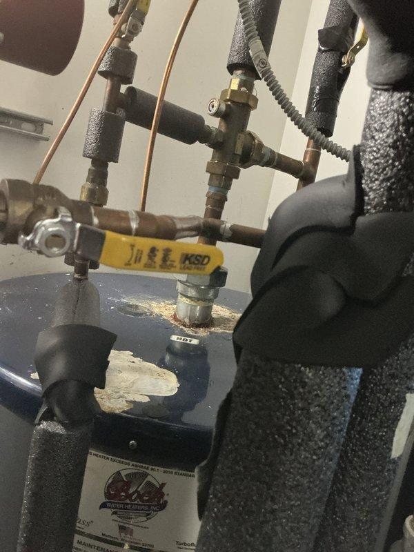 Completed water heater installation with full plumbing connections and safety components. All inlet and outlet copper piping was properly connected with brass fittings and insulated with foam pipe sleeves. Installed pressure relief valve system with discharge piping, shut-off valves with blue and yellow handles for service access, and flexible connectors for proper water flow management. Unit was secured on concrete pad with all required safety devices including temperature-pressure relief valve and drain valve at base of tank. All connections were tested and verified operational.