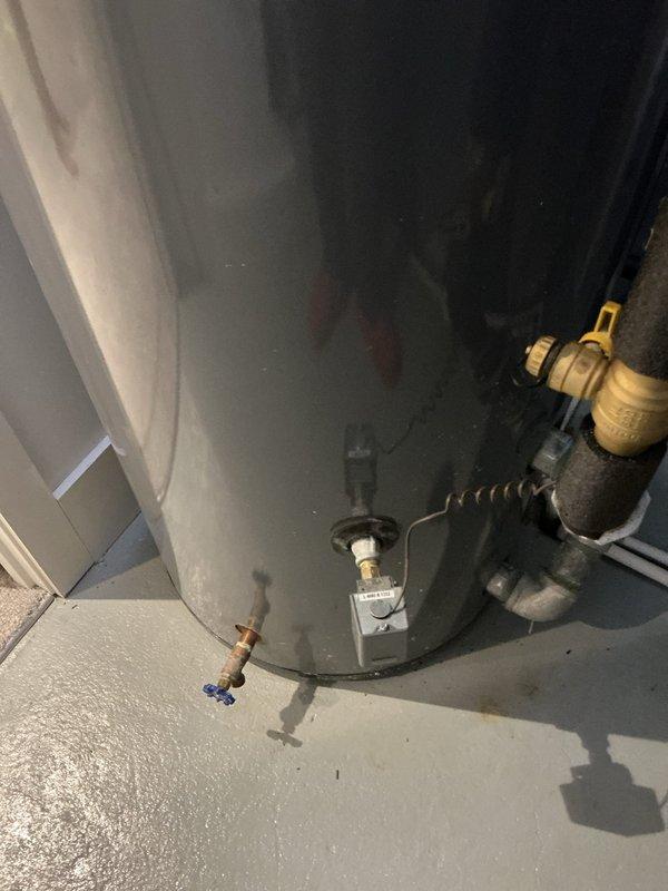 Completed water heater installation with full plumbing connections and safety components. All inlet and outlet copper piping was properly connected with brass fittings and insulated with foam pipe sleeves. Installed pressure relief valve system with discharge piping, shut-off valves with blue and yellow handles for service access, and flexible connectors for proper water flow management. Unit was secured on concrete pad with all required safety devices including temperature-pressure relief valve and drain valve at base of tank. All connections were tested and verified operational.