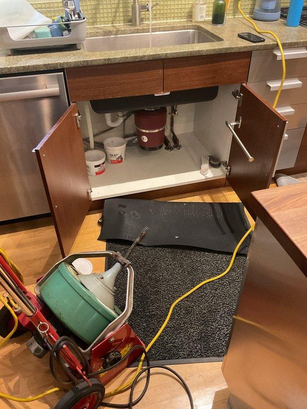 Responded to kitchen sink drainage issue where water was backing up after initially draining. Upon inspection, identified clog in the drain line beneath the sink. Deployed motorized drain cleaning machine with cable auger to clear the obstruction. Successfully removed blockage from the line, restoring proper drainage flow. Tested sink operation to confirm water now drains freely without backup. Plumbing system beneath sink, including garbage disposal and PVC drain assembly, functioning normally upon completion.
