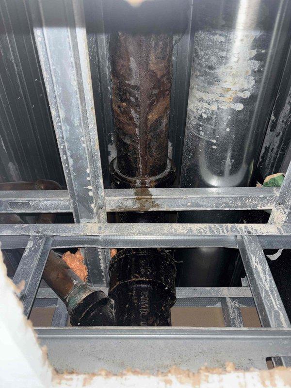 Performed water leak locate service in mechanical room area. Investigation revealed significant corrosion and rust damage on main vertical piping system, with extensive deterioration visible on bronze-colored cylinder indicating potential leak source. Documented conditions of multiple vertical pipes accessed through metal grating platform, noting heavy oxidation and moisture-related damage consistent with active or previous water intrusion. Collected physical evidence samples in containment bucket for further analysis of leak-affected materials.