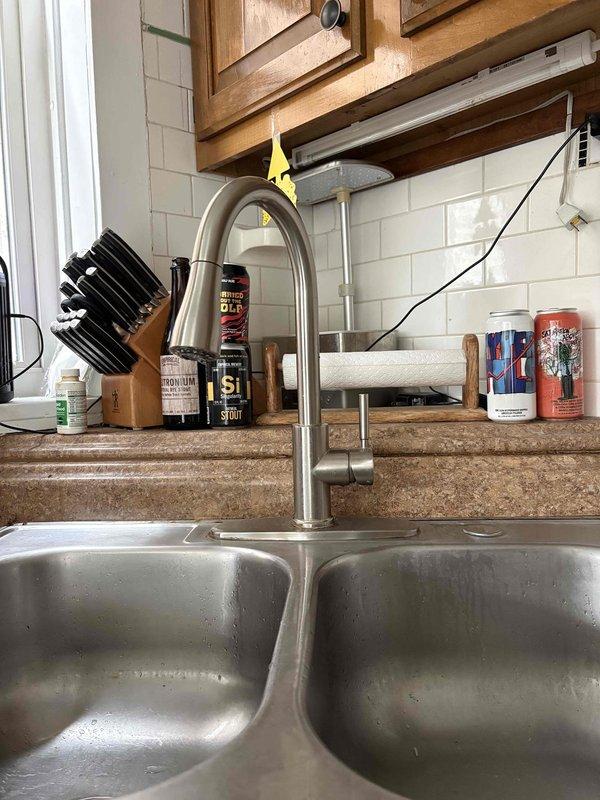 Responded to a rescheduled priority service call for a reported slow leak at the kitchen faucet. Upon arrival, conducted a thorough inspection of the kitchen sink area, including the double-basin stainless steel sink and tall brushed nickel faucet assembly. Traced water supply lines from the kitchen fixture down to the water heater connections to identify the source of the leak. Inspected flexible braided metal supply lines and connections at the water heater, examining both the hot and cold water feeds. Identified leak source and assessed condition of supply line connections, mounting hardware, and surrounding plumbing infrastructure. Documented existing conditions with photographs showing the kitchen faucet installation and the water supply routing through the wooden framing to the water heater. Completed leak location service and provided findings to customer regarding the source of the reported slow leak condition.