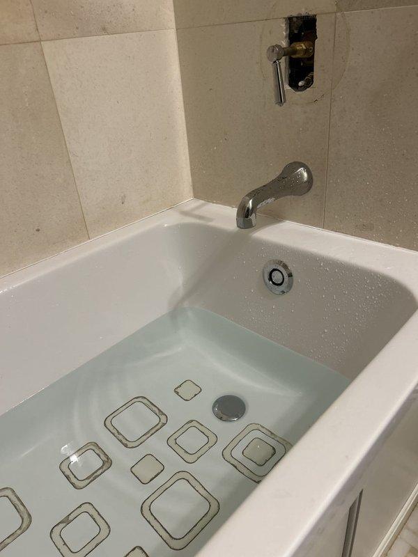 Responded to a bathtub leak affecting the unit below. Upon inspection, identified the source of water intrusion by examining the tub drain assembly and overflow connections. Accessed the plumbing system from below through the ceiling of the lower unit to assess pipe connections and identify the leak point. Inspected exposed PVC drainage piping and verified proper pitch and connections. Documented existing conditions including incomplete shower valve installation with exposed rough-in plumbing at the wall. Tightened drain connections and tested the system to confirm leak resolution.