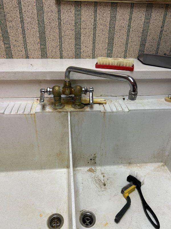 Responded to service call for non-functional faucet in utility sink. Upon arrival, inspected double-basin laundry sink with vintage-style brass valve faucet assembly and chrome spout. Diagnosed faulty valve seats and worn washers causing complete loss of water flow. Disassembled faucet valve bodies, replaced deteriorated internal components including rubber washers and brass seats on both hot and cold water supplies. Cleaned mineral deposits from valve threads and reinstalled hardware. Tested water flow and verified proper operation of both supply lines with no leaks detected. Documented existing conditions of utility room infrastructure including water meter assembly and aging plumbing connections for reference.