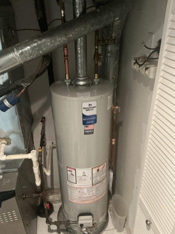 Responded to service call for leaking tanked water heater. Upon inspection, found Bradford White Defender Safety System water heater actively leaking from bottom of tank into drain pan at rapid rate, indicating tank failure. Documented condition with photographs showing the unit installed in utility closet with copper supply lines, PVC venting, and gas valve connections. Advised that tank has reached end of service life and requires replacement, as bottom leaks are not repairable due to internal tank corrosion.
