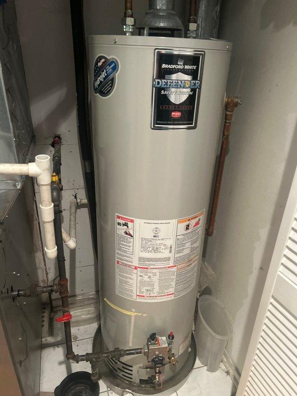 Responded to service call for leaking tanked water heater. Upon inspection, found Bradford White Defender Safety System water heater actively leaking from bottom of tank into drain pan at rapid rate, indicating tank failure. Documented condition with photographs showing the unit installed in utility closet with copper supply lines, PVC venting, and gas valve connections. Advised that tank has reached end of service life and requires replacement, as bottom leaks are not repairable due to internal tank corrosion.