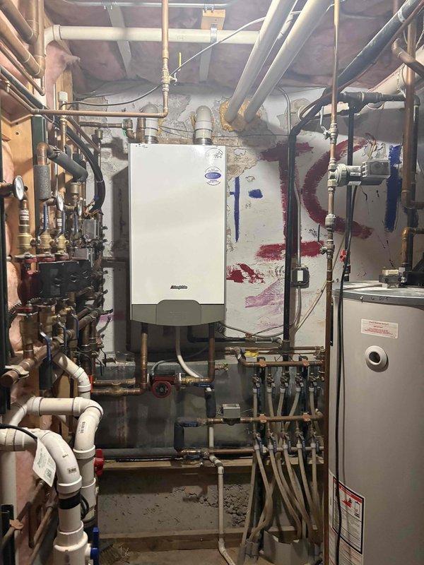Completed installation and configuration of wall-mounted tankless water heater system in basement mechanical room. Installed comprehensive copper and PVC piping network with manifold system, brass valves, and multiple branch connections to integrate new tankless unit with existing traditional storage water heater. Configured dual heating system with proper venting, pressure relief components, and shutoff valves to ensure safe and efficient operation of both heating appliances working in conjunction.