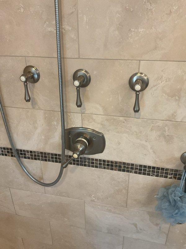 Completed installation of modern shower system featuring three-valve thermostatic control with brushed nickel lever handles and decorative mosaic tile accent border. Installed overhead rain-style showerhead with chrome finish and multiple spray patterns, along with handheld shower attachment with flexible metal hose for enhanced functionality. All fixtures were properly mounted to beige rectangular tile surfaces and tested for proper water flow and temperature control operation.