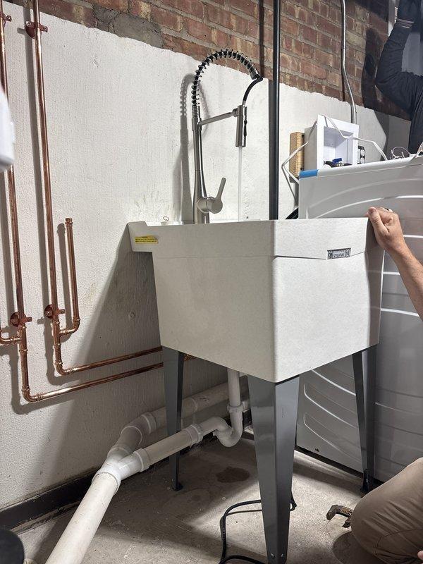 Conducted site assessment of utility sink installation in basement area to review plumbing configuration and discuss VIP membership program details with management. Inspected copper supply line routing along wall, including visible joints and fittings, verifying proper installation standards. Evaluated white PVC drainage system beneath sink, including P-trap assembly and floor-level connections. Documented existing plumbing setup featuring modern pull-down faucet with coiled spring design and chrome finish mounted on white rectangular basin with gray metal stand. Addressed questions regarding ongoing maintenance options and membership benefits for the installed plumbing fixtures.