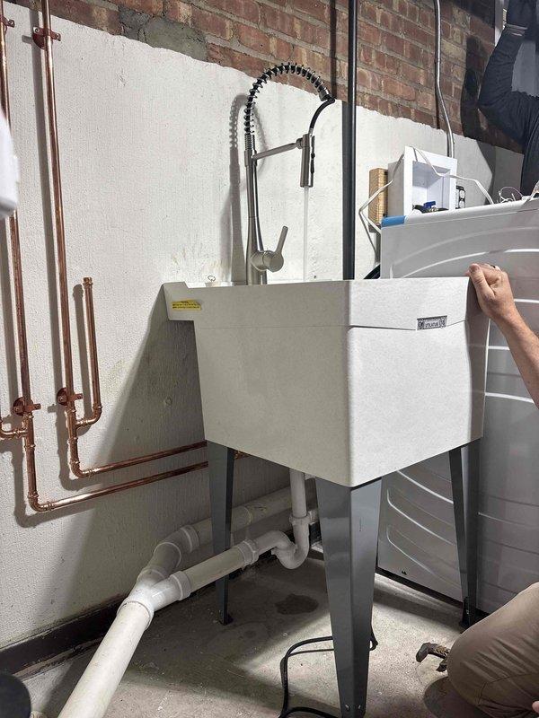 Conducted site assessment of utility sink installation in basement area to review plumbing configuration and discuss VIP membership program details with management. Inspected copper supply line routing along wall, including visible joints and fittings, verifying proper installation standards. Evaluated white PVC drainage system beneath sink, including P-trap assembly and floor-level connections. Documented existing plumbing setup featuring modern pull-down faucet with coiled spring design and chrome finish mounted on white rectangular basin with gray metal stand. Addressed questions regarding ongoing maintenance options and membership benefits for the installed plumbing fixtures.