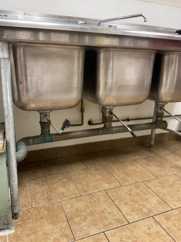 Responded to commercial kitchen for grease trap evaluation. Upon inspection, found existing 20-year-old grease trap showing significant deterioration with visible rust, corrosion around connection points, and active leak at valve body. Unit located beneath triple-basin stainless steel sink with diamond plate flooring access. Documented current condition of red metal valve assembly, inlet/outlet piping, and surrounding plumbing infrastructure. Provided assessment and recommendations for complete grease trap replacement due to age-related failure and compromised structural integrity of existing unit.
