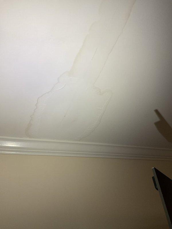 Responded to water leak originating from main bathroom and infiltrating into living room area. Upon investigation, identified active water damage evidenced by yellowish-brown discoloration and moisture staining on living room ceiling, with concentrated damage pattern along ceiling seam indicating sustained water intrusion from bathroom above. Traced leak source and documented damage extent including basement utility area where moisture penetration was affecting lower level structures. Photographed water-damaged ceiling surfaces showing linear staining patterns and discoloration, as well as basement conditions with visible floor staining near mechanical equipment and plumbing infrastructure.