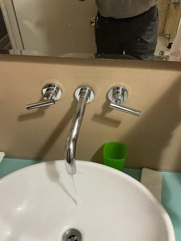 Completed installation of wall-mounted, three-piece chrome faucet assembly with center spout and dual lever handles for modern vessel sink in residential bathroom. All fixtures properly mounted, sealed, and tested for proper water flow and temperature control with no leaks detected.