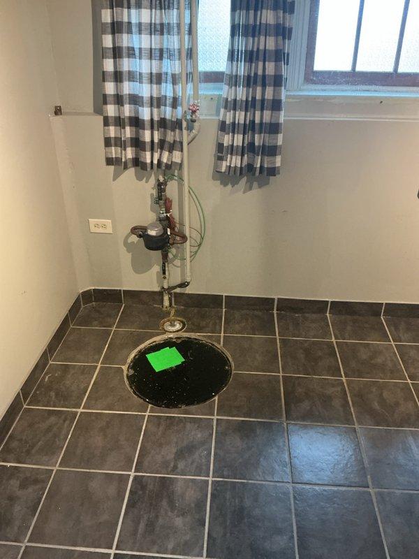 Inspected and assessed ejector pump system with visible signs of extensive wear and corrosion. The pump housing displayed significant deterioration with flaking protective coating, rusted mounting plate, and degraded gasket material indicating age-related failure. Documented existing floor drain access point and associated plumbing infrastructure including pressure gauge and valve assembly. Recommended replacement of ejector pump unit due to advanced corrosion of critical components and compromised sealing surfaces that could lead to system failure and potential flooding.
