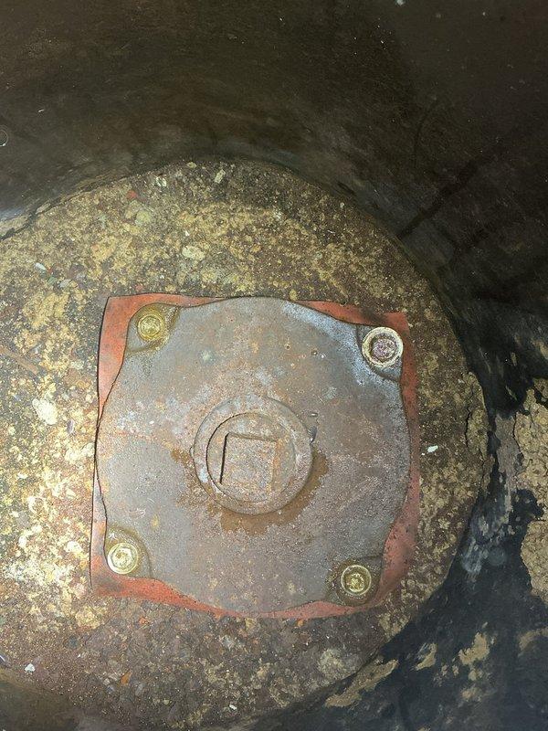 Inspected and assessed ejector pump system with visible signs of extensive wear and corrosion. The pump housing displayed significant deterioration with flaking protective coating, rusted mounting plate, and degraded gasket material indicating age-related failure. Documented existing floor drain access point and associated plumbing infrastructure including pressure gauge and valve assembly. Recommended replacement of ejector pump unit due to advanced corrosion of critical components and compromised sealing surfaces that could lead to system failure and potential flooding.