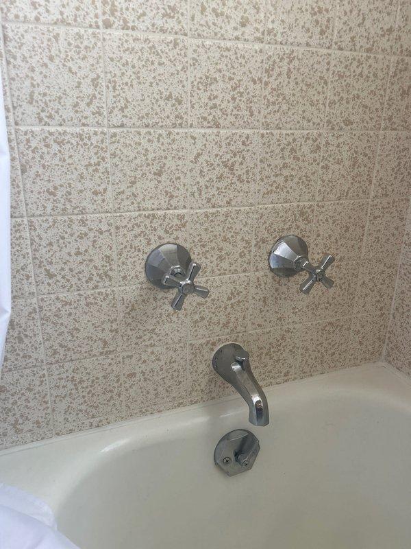 Conducted comprehensive plumbing assessment at 75-year-old residential property to provide estimates for three primary concerns. Evaluated main water valve for replacement consideration, inspecting the existing shut-off mechanism for signs of deterioration, leaking, or operational failure. Investigated second-floor bathroom shower experiencing poor water pressure issues, examining the chrome cross-handle faucet controls, tub spout, and associated supply lines beneath the fixture to identify potential causes including mineral buildup, corroded piping, or flow restrictions. Assessed two exterior hose bibs for replacement, documenting the condition of existing spigots and underlying supply connections. Photographed bathroom fixtures showing the tiled shower wall with installed chrome plumbing components and white bathtub, as well as the crawl space or basement area revealing aged, heavily rusted water supply piping with significant corrosion, mineral deposits, and deterioration consistent