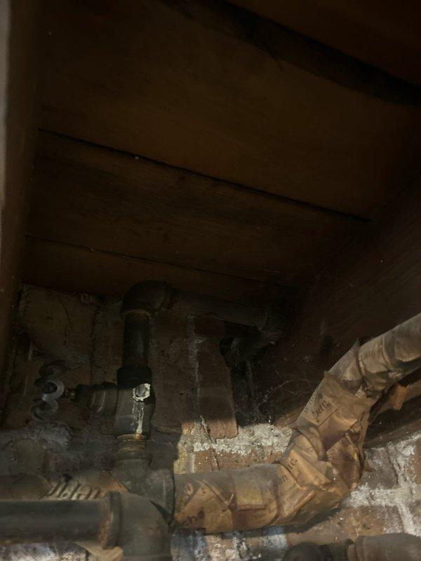 Conducted comprehensive plumbing assessment at 75-year-old residential property to provide estimates for three primary concerns. Evaluated main water valve for replacement consideration, inspecting the existing shut-off mechanism for signs of deterioration, leaking, or operational failure. Investigated second-floor bathroom shower experiencing poor water pressure issues, examining the chrome cross-handle faucet controls, tub spout, and associated supply lines beneath the fixture to identify potential causes including mineral buildup, corroded piping, or flow restrictions. Assessed two exterior hose bibs for replacement, documenting the condition of existing spigots and underlying supply connections. Photographed bathroom fixtures showing the tiled shower wall with installed chrome plumbing components and white bathtub, as well as the crawl space or basement area revealing aged, heavily rusted water supply piping with significant corrosion, mineral deposits, and deterioration consistent