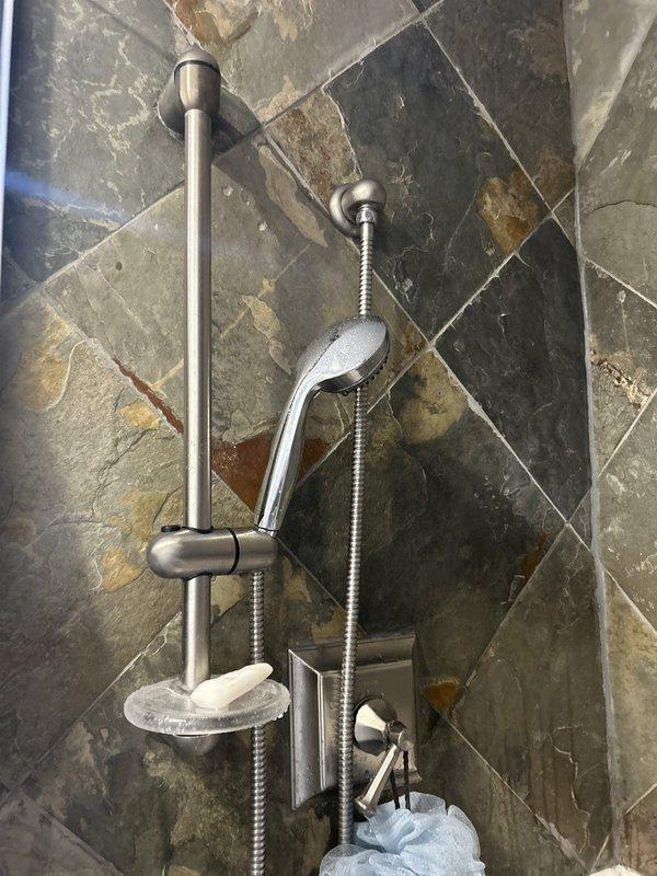 Responded to service call for leaking shower hose in residential bathroom. Upon inspection, assessed handheld shower assembly mounted on chrome slide bar rail system installed over diagonal slate tile wall. Examined flexible metal hose connection points at both the wall-mounted diverter valve and handheld shower head fixture for signs of wear, deterioration, or improper sealing. Evaluated hose integrity along entire length and tested connections under water pressure to identify source of leak. Inspected mounting hardware, slide bar brackets, and wall-mounted control valve for proper installation and function. Documented existing conditions of shower fixture assembly and tile surround, noting natural mineral deposits and water staining typical of slate material in wet environment. Photographed shower head, flexible hose, rail system, and surrounding tilework for service records.