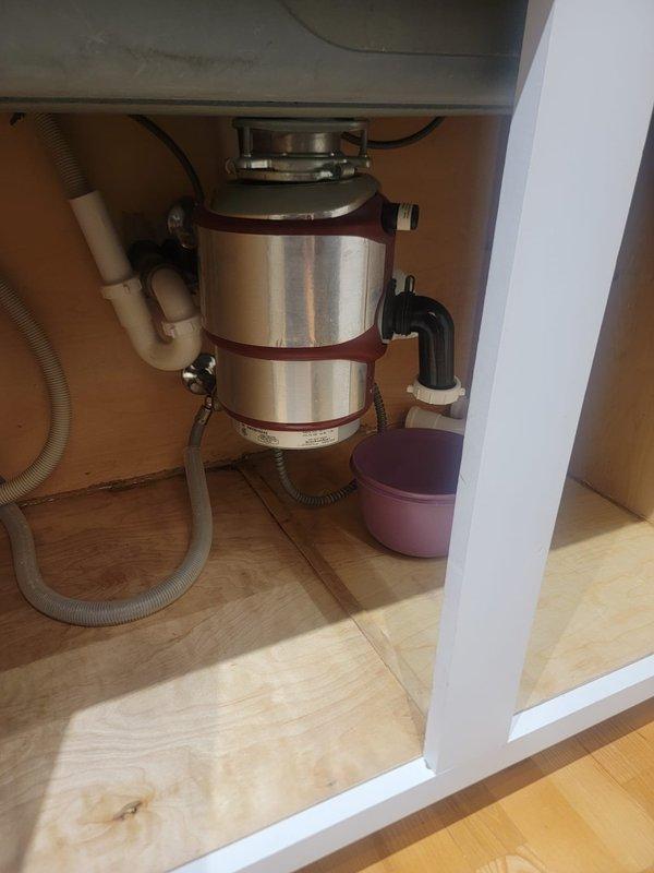 Responded to a clogged kitchen sink complaint and performed diagnostic inspection of the drainage system. Upon examination of the under-sink plumbing configuration, assessed the garbage disposal unit, P-trap assembly, and associated drain lines for blockages or mechanical failures. Documented existing plumbing conditions including the disposal unit with rubber splash guard, PVC drainage piping, corrugated drain hose connections, and trap configuration to identify the source of the stoppage and determine appropriate clearing methods.