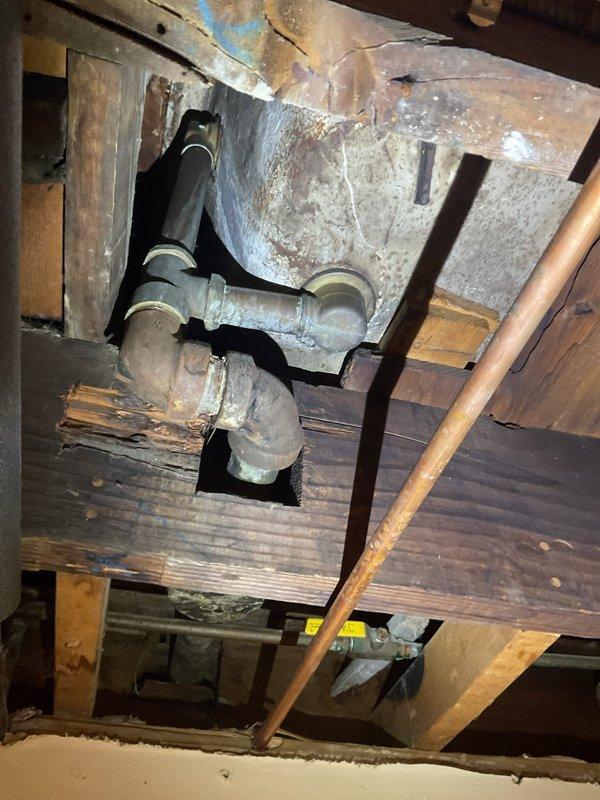Responded to water leak in basement originating from exposed plumbing in ceiling below bathroom. Upon inspection, identified active leak from PVC pipe junction in floor framing system with clean water supply line affected. Documented existing plumbing configuration including PVC T-junction, copper supply lines, and cast iron components routed through floor joists and subfloor structure. Assessed leak source and determined water intrusion was from potable water supply system rather than wastewater drainage. Photographed affected plumbing connections and surrounding structural elements for documentation and further repair planning.