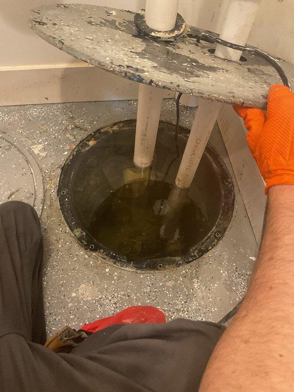 Responded to urgent callback for wet basement floor and non-functioning sump pump following previous check valve replacement. Upon arrival, inspected sump pit and found pump inoperative with moisture present on cap assembly. Observed standing water in basement pit with PVC discharge piping submerged in murky water. Assessed pump functionality and verified check valve operation from prior service visit. Documented conditions showing deteriorated wooden pit cover and water accumulation requiring immediate attention to prevent further basement flooding.