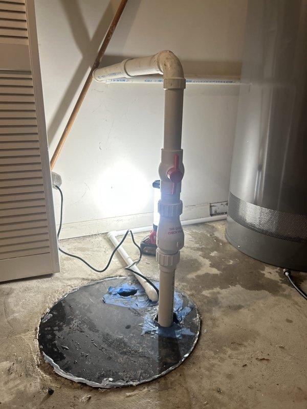 Responded to service call for sump pump running continuously in basement utility area. Upon inspection, found existing sump pump system with PVC discharge piping and check valve assembly. Assessed pump operation and basin conditions using work light to examine interior components. Identified pump cycling issues causing continuous runtime. Evaluated discharge pipe configuration, check valve functionality, and float switch operation. Inspected electrical connections and pump motor performance. Documented existing installation showing vertical PVC discharge assembly with elbow joint, concrete basin seal, and associated plumbing components for customer records and replacement recommendations.