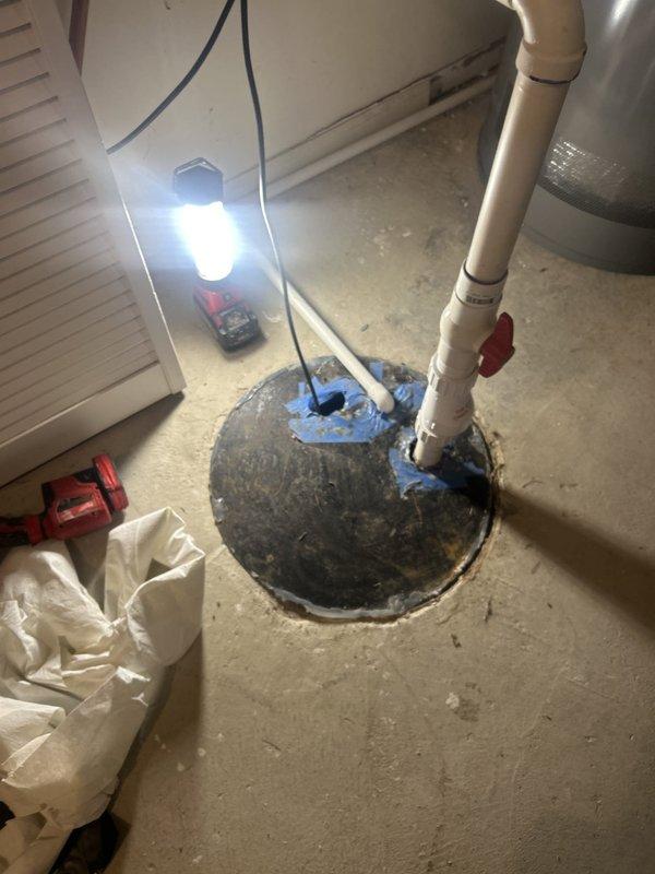 Responded to service call for sump pump running continuously in basement utility area. Upon inspection, found existing sump pump system with PVC discharge piping and check valve assembly. Assessed pump operation and basin conditions using work light to examine interior components. Identified pump cycling issues causing continuous runtime. Evaluated discharge pipe configuration, check valve functionality, and float switch operation. Inspected electrical connections and pump motor performance. Documented existing installation showing vertical PVC discharge assembly with elbow joint, concrete basin seal, and associated plumbing components for customer records and replacement recommendations.