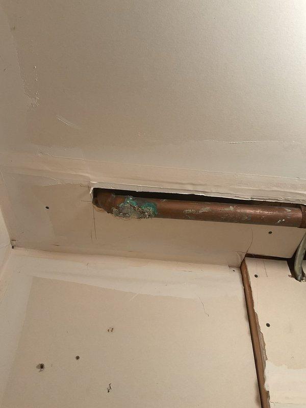 Accessed corroded copper supply piping through wall opening in bathroom area and exposed PVC drain lines through ceiling access point to assess plumbing system condition. Documented significant oxidation and patina buildup on horizontal copper pipe run, indicating active corrosion requiring replacement. Removed wall sections and access panels to expose drainage infrastructure including white PVC waste lines with multiple fittings and connections. Identified aged copper piping behind PVC system requiring remediation to prevent future leaks and water damage.