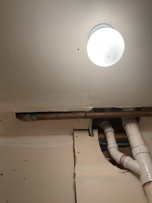 Accessed corroded copper supply piping through wall opening in bathroom area and exposed PVC drain lines through ceiling access point to assess plumbing system condition. Documented significant oxidation and patina buildup on horizontal copper pipe run, indicating active corrosion requiring replacement. Removed wall sections and access panels to expose drainage infrastructure including white PVC waste lines with multiple fittings and connections. Identified aged copper piping behind PVC system requiring remediation to prevent future leaks and water damage.