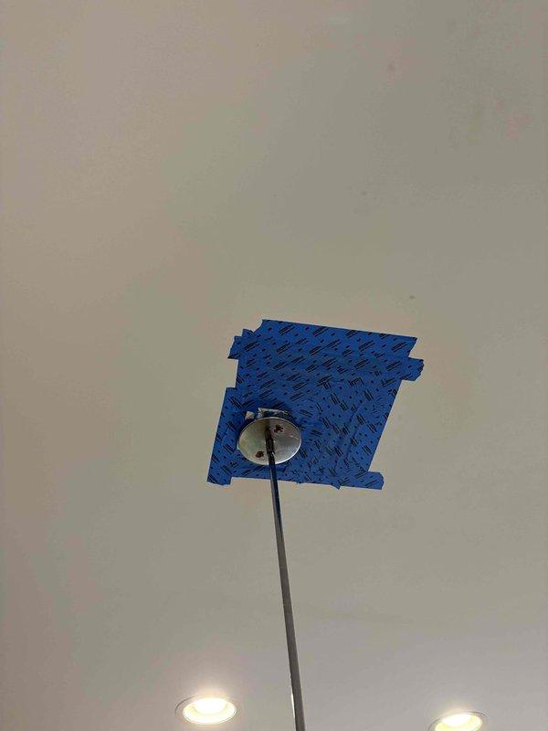 Responded to plumbing service call for ceiling-mounted light fixture installation or repair. Upon arrival, accessed ceiling penetration point where fixture pole extends through drywall. Masked off work area with painter's tape in crosshatch pattern to protect surrounding ceiling surface during repair work. Secured pendant light fixture with chrome mounting canopy at ceiling junction, ensuring proper alignment and stability of extended pole assembly. Completed installation with fixture properly mounted and operational alongside existing recessed lighting.