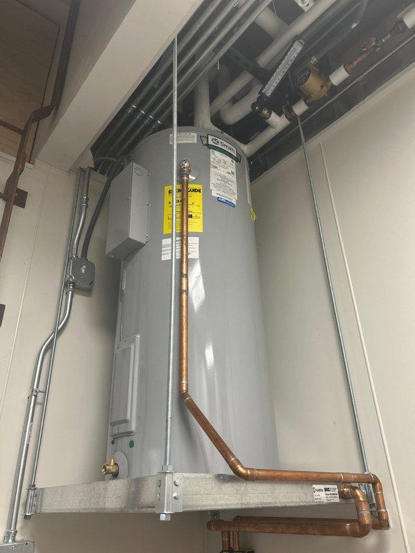 Completed commercial water heater estimate for replacement of existing tall cylindrical unit in utility room. Assessed current installation including copper supply and discharge piping connections, electrical conduit routing to junction box, and proper venting through ceiling cavity. Documented water heater mounted on elevated white metal platform with multiple building systems including HVAC ductwork, gray PVC piping, and various conduits running overhead in exposed ceiling space.