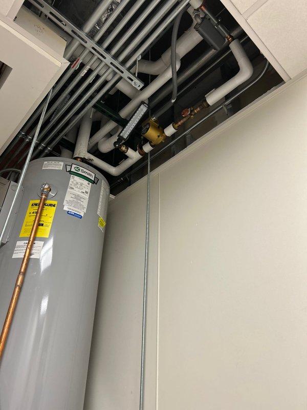 Completed commercial water heater estimate for replacement of existing tall cylindrical unit in utility room. Assessed current installation including copper supply and discharge piping connections, electrical conduit routing to junction box, and proper venting through ceiling cavity. Documented water heater mounted on elevated white metal platform with multiple building systems including HVAC ductwork, gray PVC piping, and various conduits running overhead in exposed ceiling space.