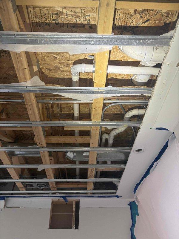 # Plumbing Inspection - Exposed Ceiling Cavity

Responded to service call for inspection of plumbing system after customer opened ceiling to investigate previous concerns. Conducted thorough visual assessment of exposed ceiling cavity containing multiple building systems including white PVC drain and vent piping, HVAC ductwork, and structural framing components.

Examined PVC plumbing configuration running through wooden joists and OSB subfloor, noting pipe routing, elbow joints, mounting hardware, and penetrations through framing members. Inspected integration of plumbing system with galvanized metal HVAC ducts and cross-bracing support members within the ceiling space. Documented existing conditions with photographs showing complete layout of drainage system, structural framework, and mechanical systems for assessment and estimate preparation. Advised customer on findings and next steps for necessary repairs.