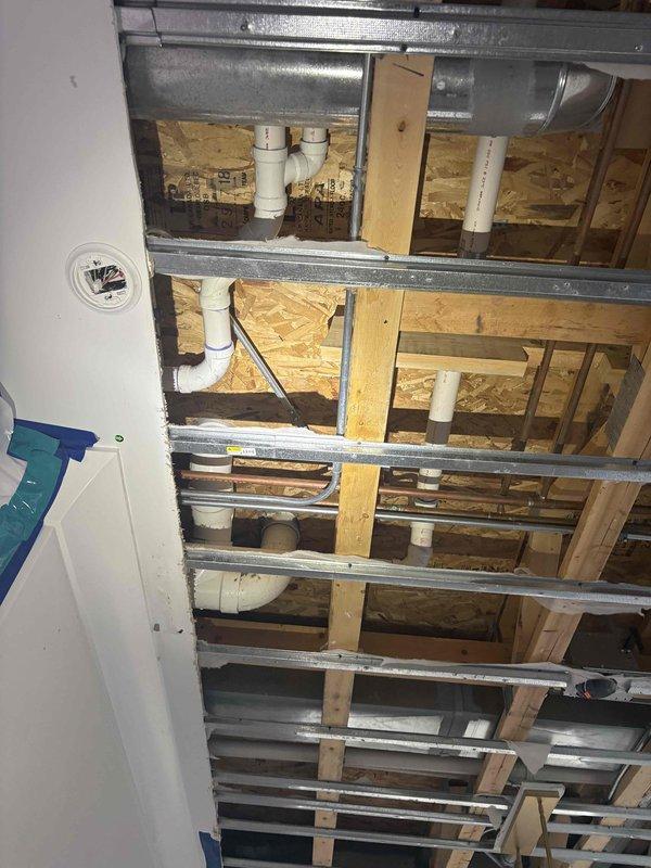 # Plumbing Inspection - Exposed Ceiling Cavity

Responded to service call for inspection of plumbing system after customer opened ceiling to investigate previous concerns. Conducted thorough visual assessment of exposed ceiling cavity containing multiple building systems including white PVC drain and vent piping, HVAC ductwork, and structural framing components.

Examined PVC plumbing configuration running through wooden joists and OSB subfloor, noting pipe routing, elbow joints, mounting hardware, and penetrations through framing members. Inspected integration of plumbing system with galvanized metal HVAC ducts and cross-bracing support members within the ceiling space. Documented existing conditions with photographs showing complete layout of drainage system, structural framework, and mechanical systems for assessment and estimate preparation. Advised customer on findings and next steps for necessary repairs.