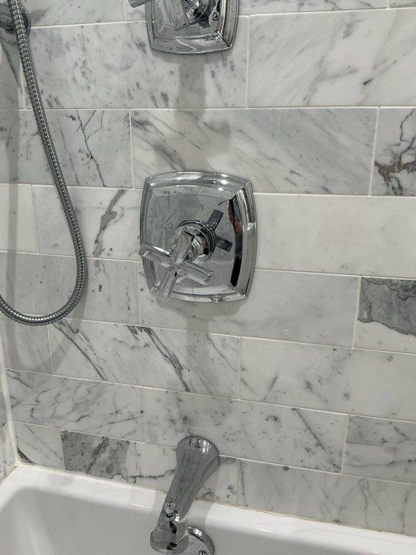 Completed installation of new shower fixtures in bathroom including chrome shower valve with cross-handle control, showerhead, handheld shower with flexible hose, and tub spout. All fixtures were mounted on newly installed white and gray marble-patterned tile work, with proper sealing and alignment to ensure leak-free operation.