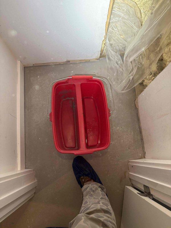 Responded to report of water leak in basement ceiling near main water supply entry point. Upon investigation, accessed ceiling cavity and located active leak at copper plumbing assembly where main water line enters the structure. Identified compromised solder joint on copper pipe fitting as source of water intrusion. Placed collection bucket to contain dripping water and prevent further damage to basement floor and surrounding areas. Documented conditions including exposed insulation, plumbing configuration with copper pipes and white cylindrical fixture, and extent of moisture penetration in the affected area for repair recommendations.