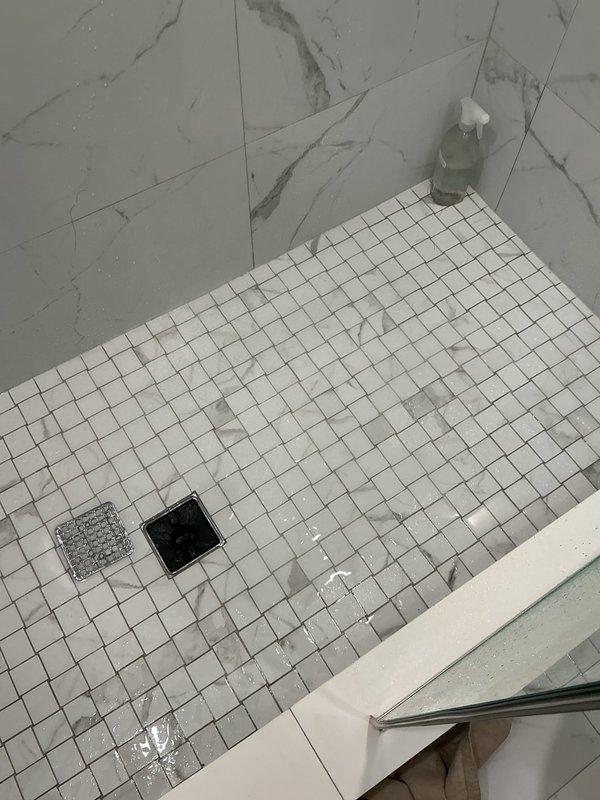 Responded to clogged shower drain for new customer referred by HOA president. Upon arrival, assessed shower stall with white mosaic tile floor and dual drain configuration. Identified slow drainage issue in primary shower drain. Utilized plunger to clear blockage, working the obstruction through the drain line. Successfully restored proper water flow and drainage function. Tested system to confirm complete clearance. Captured documentation photos showing before and after conditions of the shower drain area.