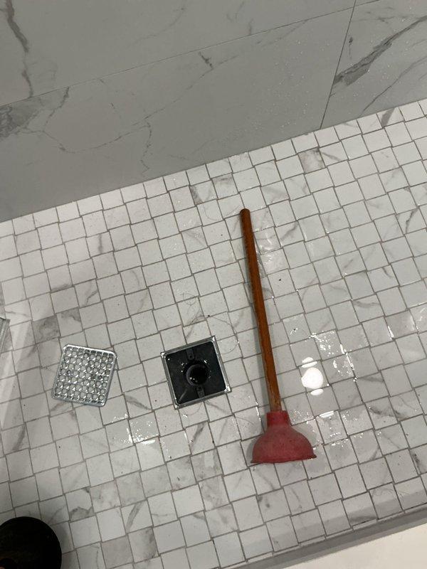 Responded to clogged shower drain for new customer referred by HOA president. Upon arrival, assessed shower stall with white mosaic tile floor and dual drain configuration. Identified slow drainage issue in primary shower drain. Utilized plunger to clear blockage, working the obstruction through the drain line. Successfully restored proper water flow and drainage function. Tested system to confirm complete clearance. Captured documentation photos showing before and after conditions of the shower drain area.