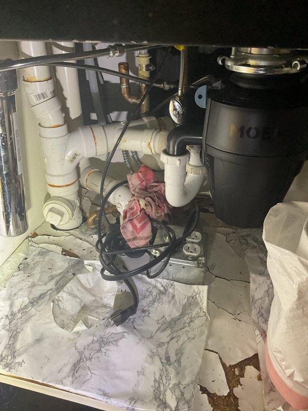Responded to kitchen sink clog and backup issue. Upon arrival, accessed the under-sink cabinet area and inspected the drain assembly, which included white PVC piping, multiple joints and connections, and a black garbage disposal unit. Disassembled drain components to locate and clear the obstruction causing the backup. Removed accumulated debris from the drain line and verified proper flow through the system. Reassembled all plumbing connections, tested the disposal unit operation, and confirmed drainage was functioning properly with no leaks present at any connection points.