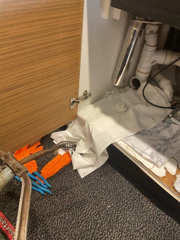 Responded to kitchen sink clog and backup issue. Upon arrival, accessed the under-sink cabinet area and inspected the drain assembly, which included white PVC piping, multiple joints and connections, and a black garbage disposal unit. Disassembled drain components to locate and clear the obstruction causing the backup. Removed accumulated debris from the drain line and verified proper flow through the system. Reassembled all plumbing connections, tested the disposal unit operation, and confirmed drainage was functioning properly with no leaks present at any connection points.