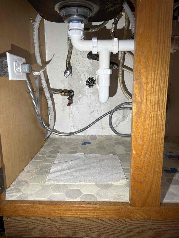 Responded to warranty callback for kitchen stack leak affecting multiple floors. Upon inspection, found water accumulation under sink cabinet following previous service visit. Inspected PVC drain assembly, P-trap configuration, garbage disposal connections, and all slip-joint fittings for proper sealing and alignment. Documented existing plumbing configuration including white PVC trap assembly, supply lines, shut-off valves, and disposal unit connections to verify installation integrity and identify source of continued leakage.